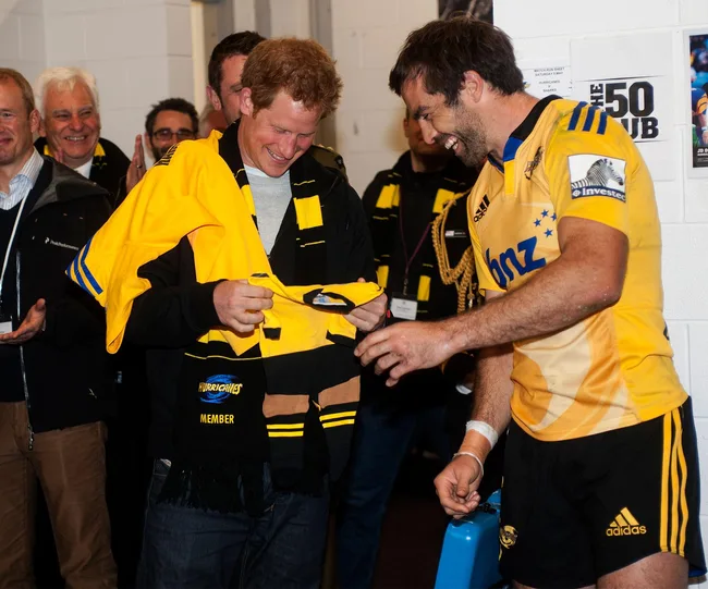 Two men smiling, one in a yellow rugby jersey and the other holding a yellow team shirt indoors.