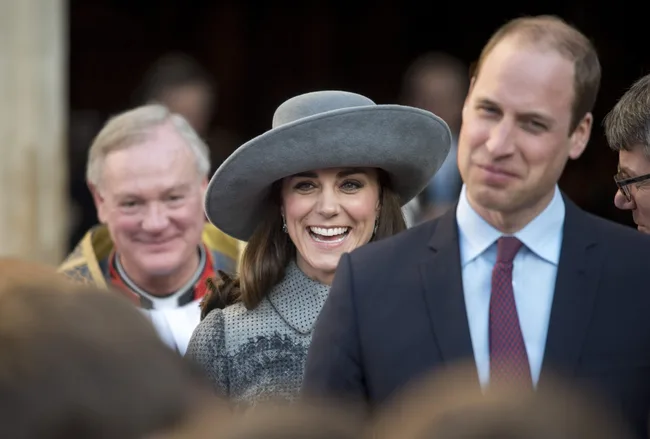 Smiling woman in a broad gray hat and man in a suit, standing outdoors with a crowd.