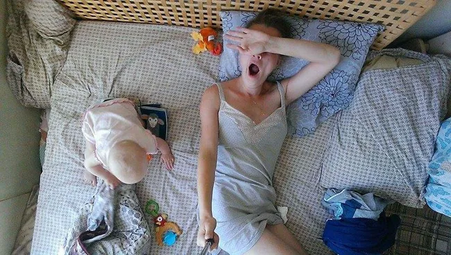 Mother and baby on bed; mother yawns while taking a selfie, baby plays with a toy.