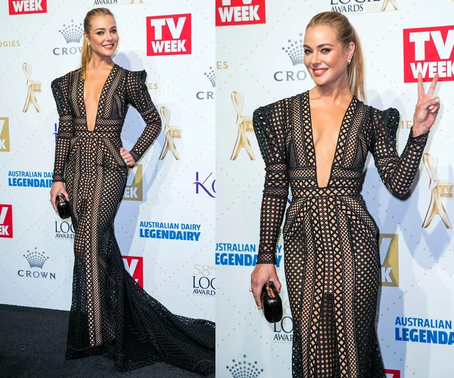 A woman in a black, patterned dress at an awards event, posing on the red carpet with a smile and peace sign.