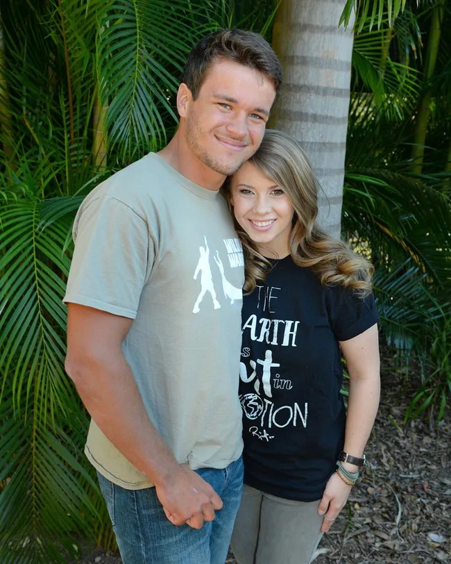 A smiling couple stands closely together in front of tropical plants. Both wear casual t-shirts.
