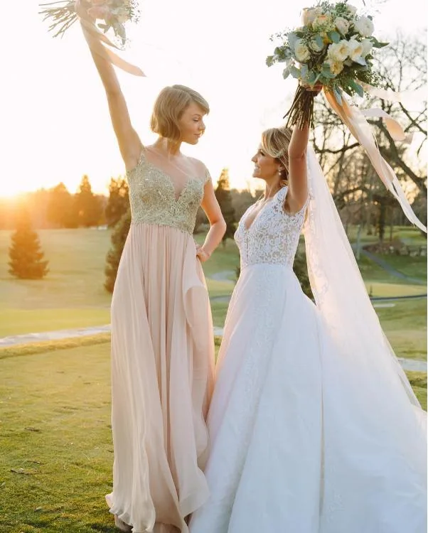 Women in elegant dresses hold bouquets and raise arms joyfully on a sunny outdoor occasion.