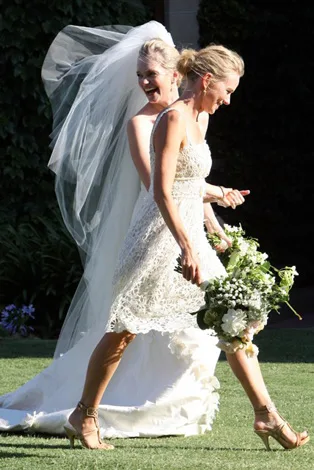 Bride in white gown and veil joyfully walks with bridesmaid holding bouquet on sunny lawn.