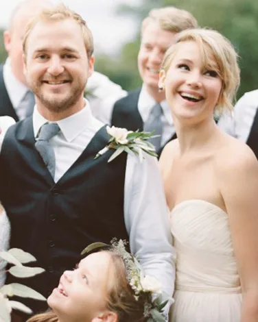 A couple smiling at a wedding, with greenery in the background and a child looking up at them.