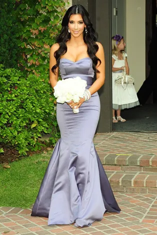 Brunette woman in a strapless lavender gown holding a white bouquet, standing on a walkway with greenery and a flower girl nearby.