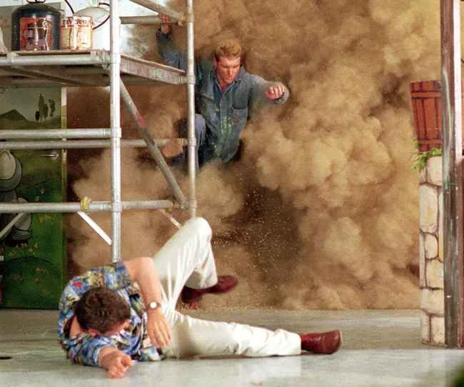 Two men in chaotic scene with dust cloud; one falling, one jumping from scaffolding indoors.