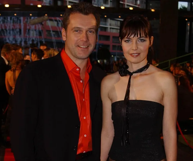 A man in a black suit with a red shirt and a woman in a black strapless dress pose together at a formal event.