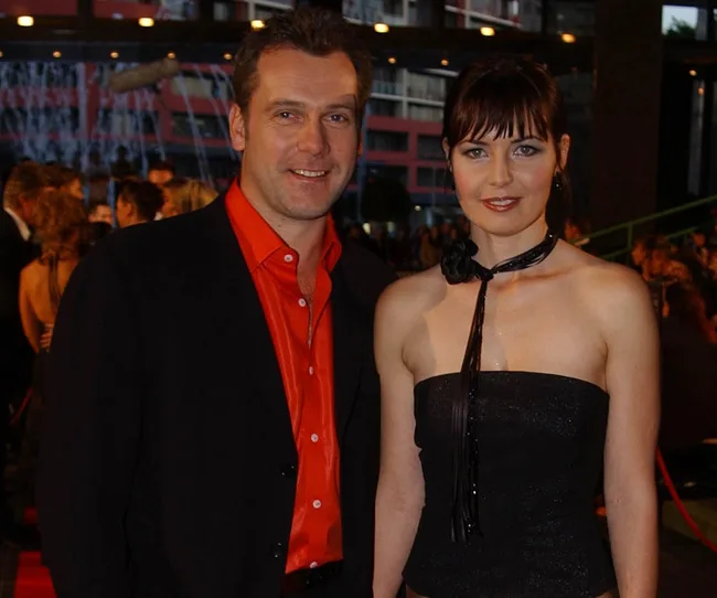 A man in a black suit with a red shirt and a woman in a black strapless dress pose together at a formal event.
