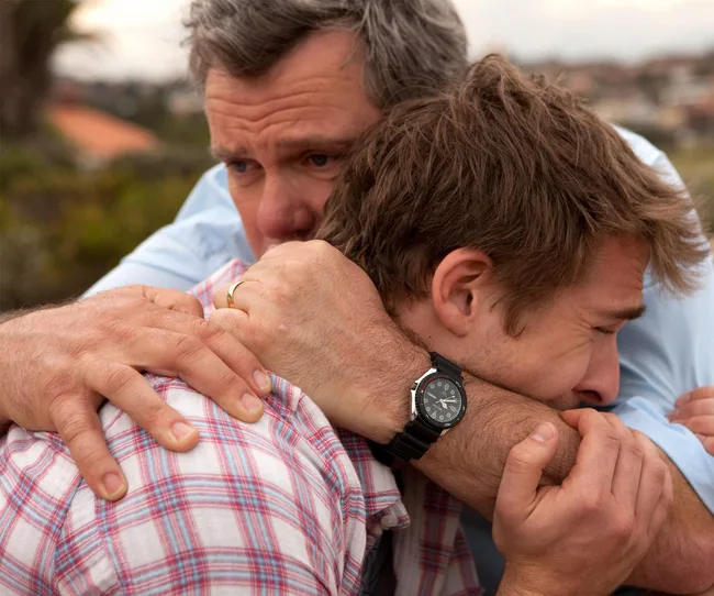 A man in a plaid shirt is being comforted by an older man wearing a watch, embracing him gently.