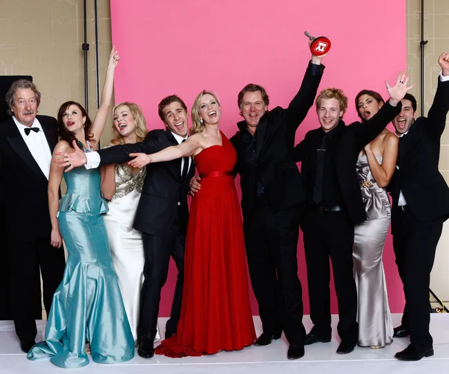 Group of nine people in formal attire celebrating in front of a pink backdrop, one holding an award.