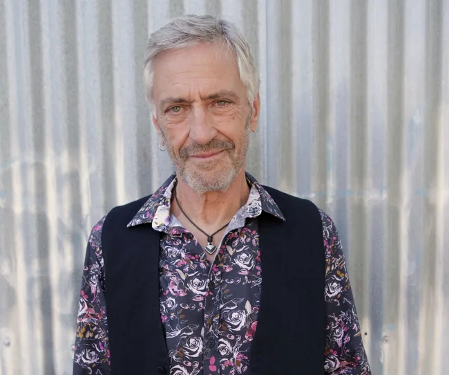 Man with short gray hair, wearing a floral shirt and black vest, stands against a corrugated metal background.