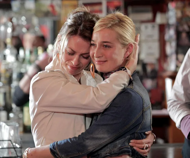 Two women embrace emotionally in a bar; one appears to be comforting the other.