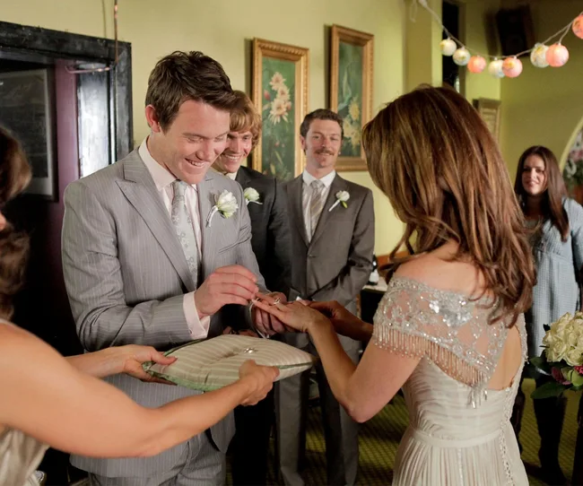 A bride and groom exchange rings at a wedding, surrounded by smiling guests.