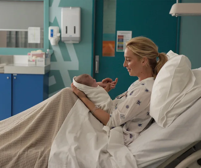 A woman lies in a hospital bed smiling at a newborn baby wrapped in a blanket.
