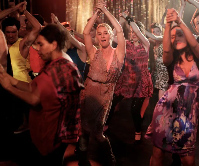 Group of people dancing energetically in a vibrant, dimly lit setting with colorful dresses and background decorations.