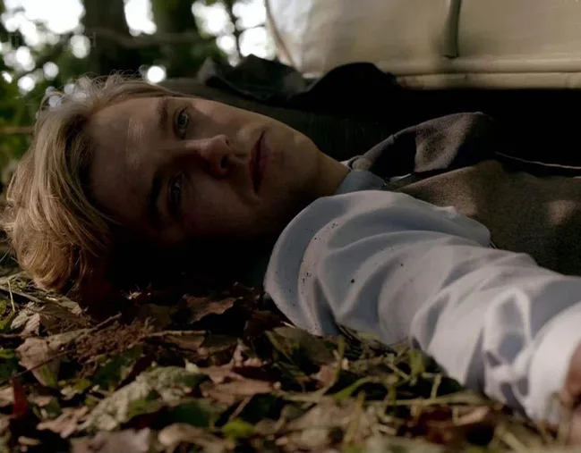 A man in a white shirt lies on the ground outdoors, gazing upwards with a contemplative expression.