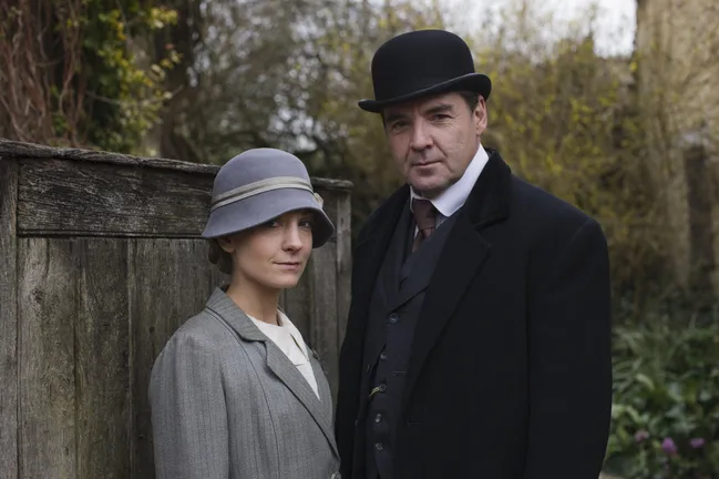 Man and woman in early 20th-century attire, standing outdoors by a wooden fence with foliage in the background.
