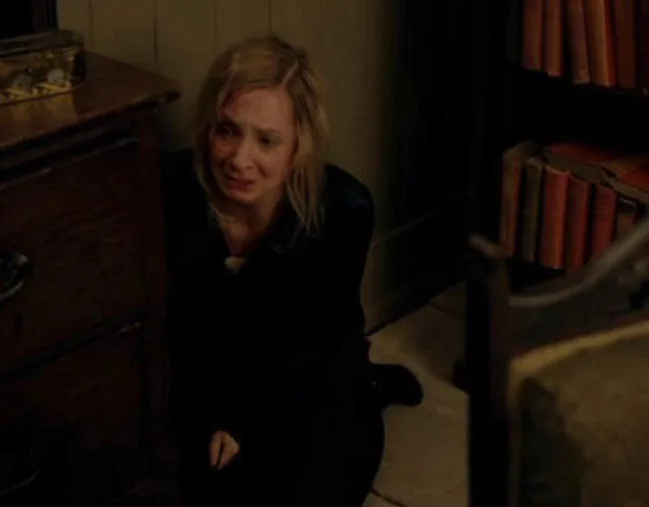 A distressed woman sits on the floor beside a wooden chest and bookshelf, tears in her eyes.