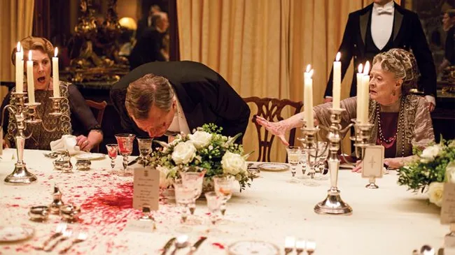 A man leans over a lavish dining table, spilling wine, as two women react in shock amidst ornate tableware and candles.