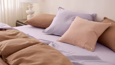 Bed with lavender sheets, tan pillows, and a white lamp on a nightstand.