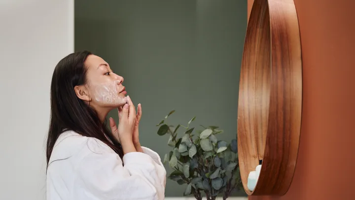 Woman in a white robe applying face cream while looking in a wooden-frame mirror in a bathroom setting.