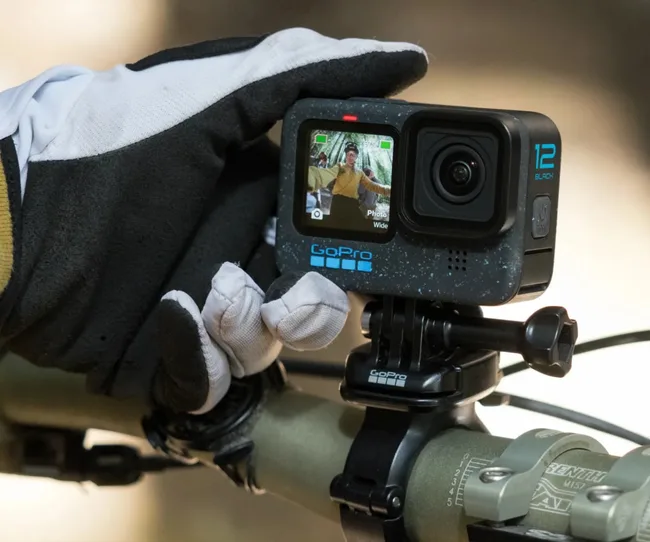 A gloved hand adjusts a GoPro Hero 12 Black camera mounted on a bicycle handlebar.