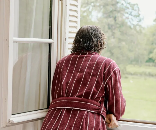 Person in a striped robe leans out of a window, looking at a green landscape.