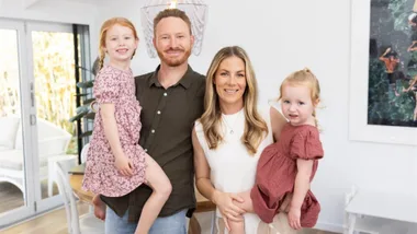 A family of four smiling in a bright living room, with the parents holding two young daughters.