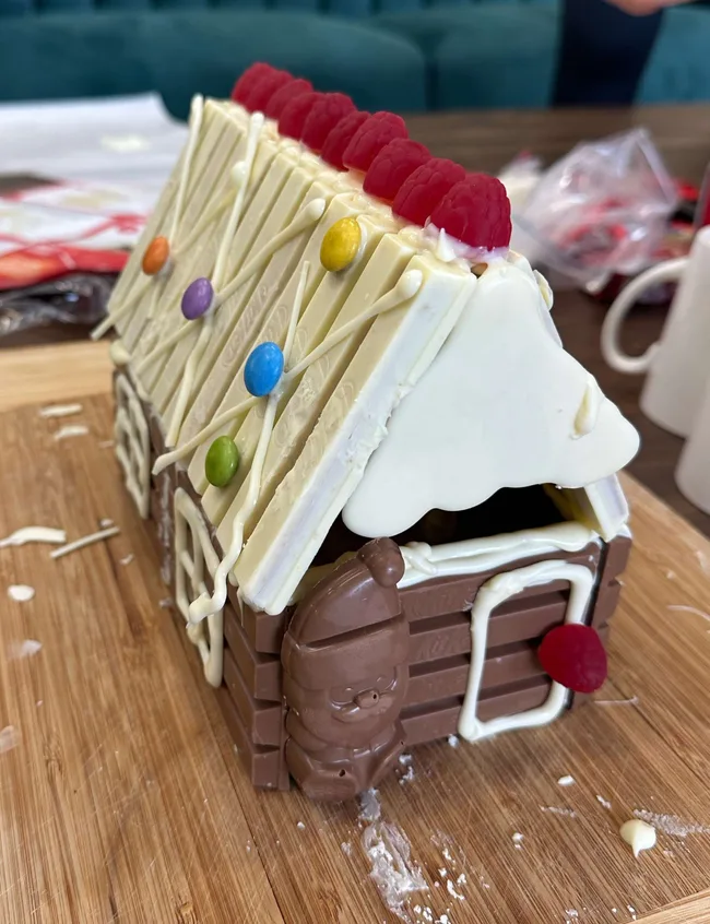 A chocolate gingerbread house decorated with colored candies and gummy raspberries on a wooden board.