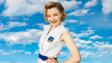 A woman in a vintage-style white outfit smiles against a blue sky with clouds.
