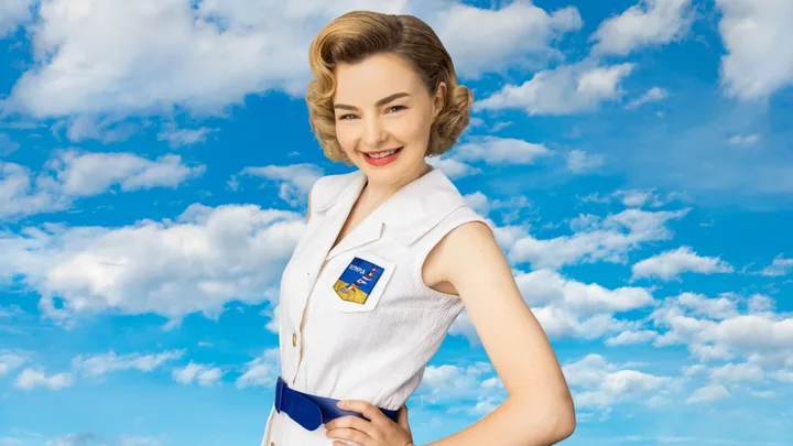 A woman in a vintage-style white outfit smiles against a blue sky with clouds.