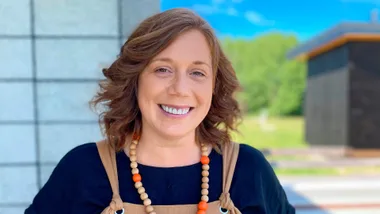A woman with shoulder-length brown hair and a beaded necklace smiles outdoors.