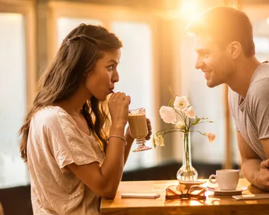 Man and woman sharing a drink at a cafe, smiling at each other, with sunlight streaming in.