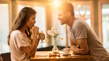 Man and woman sharing a drink at a cafe, smiling at each other, with sunlight streaming in.