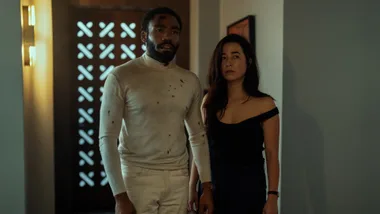 A man in a blood-stained white shirt stands beside a woman in a black top in a dimly lit hallway.