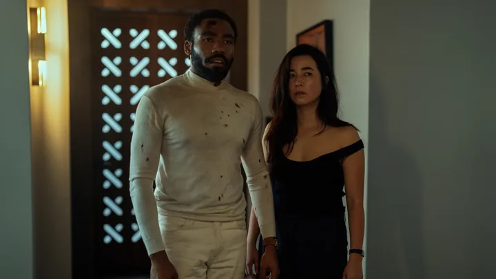 A man in a blood-stained white shirt stands beside a woman in a black top in a dimly lit hallway.