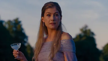 A woman in a shimmering dress holds a cocktail against a blurred outdoor background under evening light.
