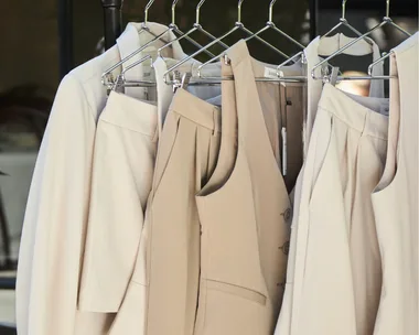 Person in black blazer with coffee cup on left; beige suits on hangers on right.