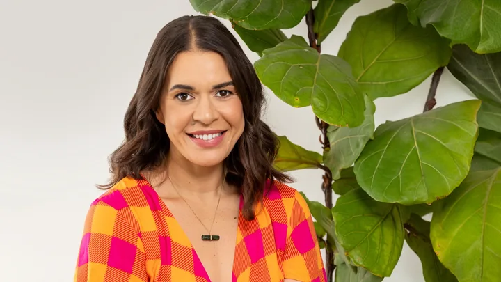 Smiling woman in a colorful checkered top stands beside a lush green plant.