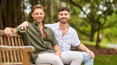 Two men sitting on a park bench, smiling, one wearing a green shirt, the other in white; lush greenery in the background.