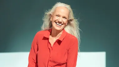 Elderly woman with long white hair, smiling, wearing a red blouse against a teal background.