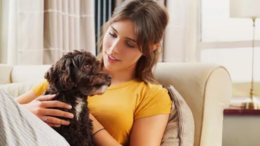 A woman in a yellow shirt lovingly holds a small, fluffy dog on a couch in a cozy living room.
