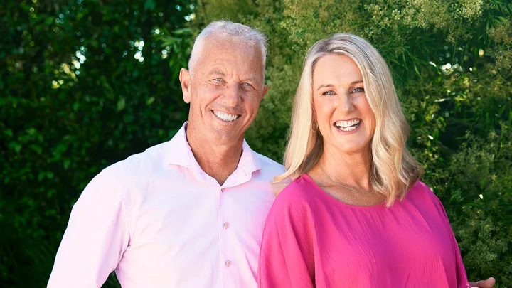 Man in light pink shirt and woman in fuchsia blouse smiling outdoors with greenery in the background.