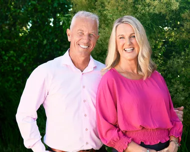 Man in light pink shirt and woman in fuchsia blouse smiling outdoors with greenery in the background.