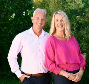 Man in light pink shirt and woman in fuchsia blouse smiling outdoors with greenery in the background.