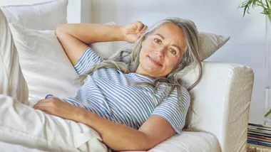 Senior woman with gray hair in a striped shirt relaxing on a white couch, smiling gently.