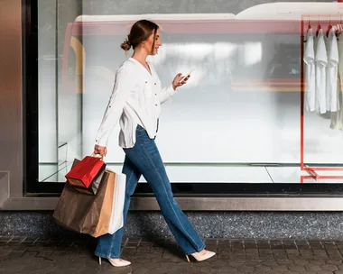 Woman walking with shopping bags, wearing jeans and a white shirt, looking at her phone outside a store with white shirts displayed.