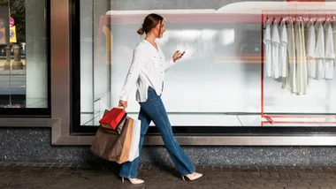 Woman walking with shopping bags, wearing jeans and a white shirt, looking at her phone outside a store with white shirts displayed.