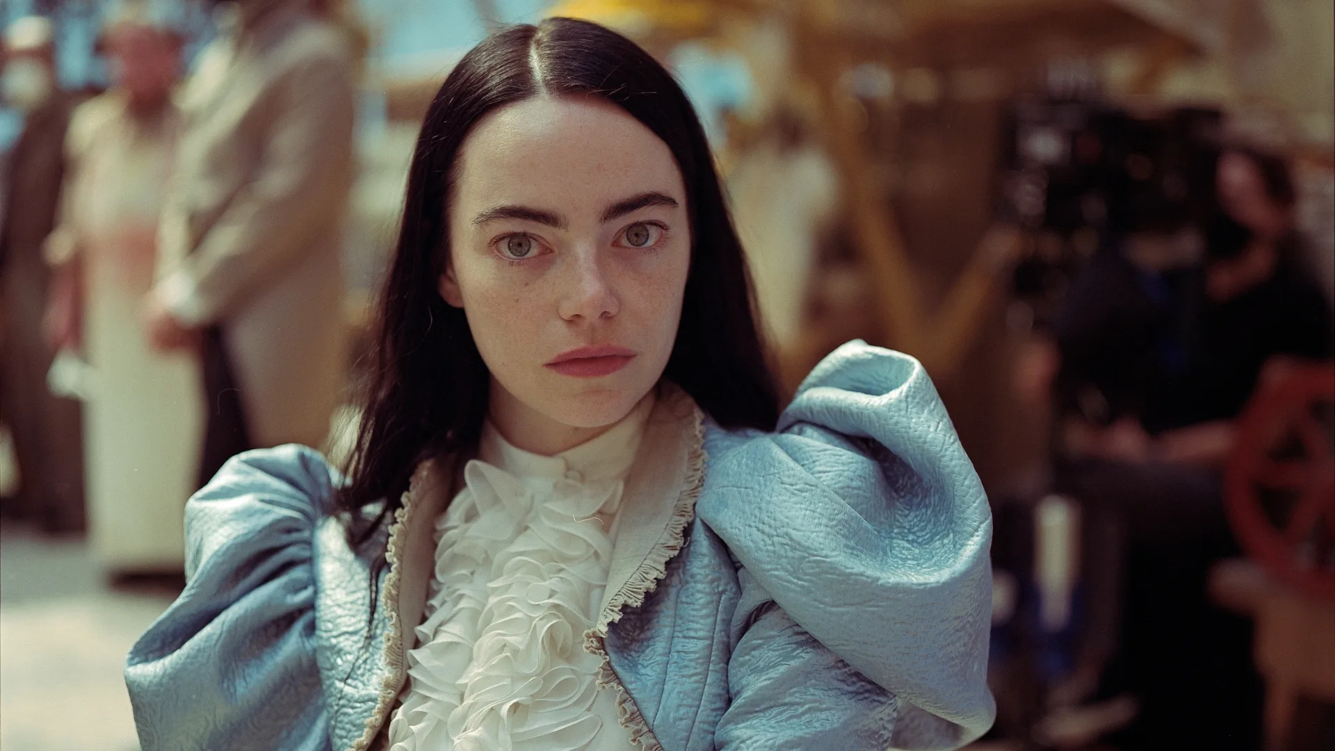 A woman with long dark hair in a blue costume looks seriously at the camera, blurred background with people.