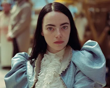 A woman with long dark hair in a blue costume looks seriously at the camera, blurred background with people.